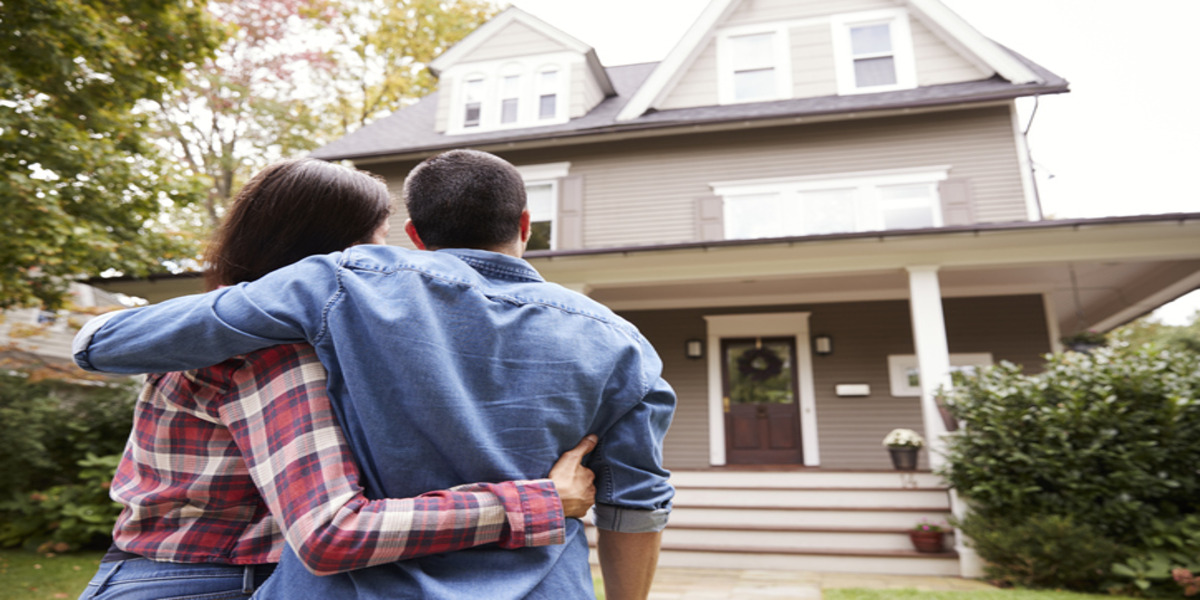 A couple standing close together with their backs to the camera, embracing while looking at a two-story house with a front porch and a main entrance adorned with a wreath. It's a residential setting with overcast skies, suggesting a calm, possibly autumnal day.