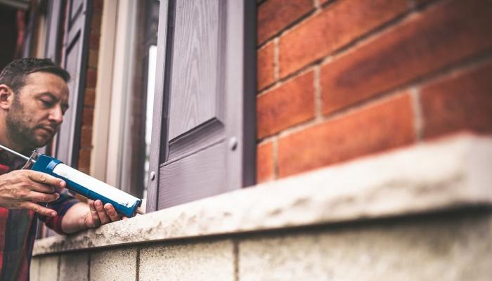 A man applies caulk to the window of a brick home.