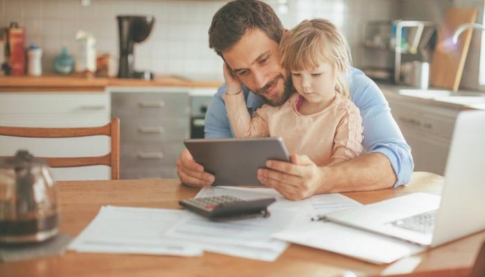 A father and toddler look at a tablet while doing paperwork at the kitchen table.