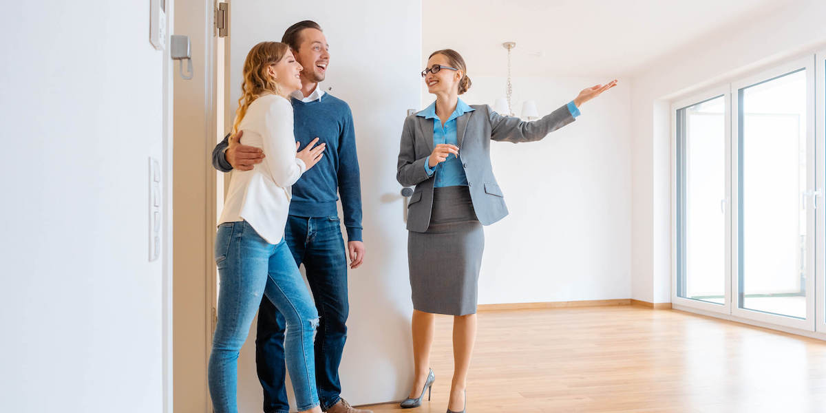 Real estate agent showing a spacious room to a happy couple in a house tour, with the agent gesturing towards large windows with natural light.