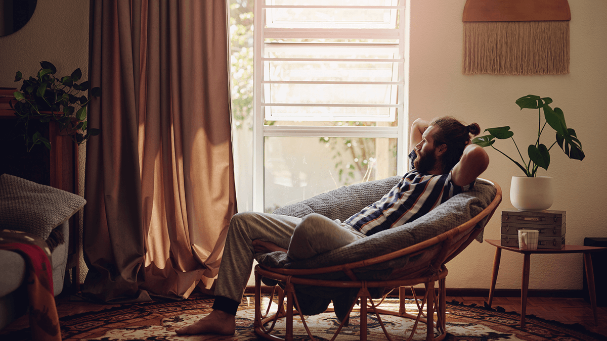 Man reclines in a shell shaped chair while looking out the window of his home.