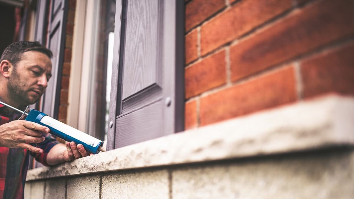 A man applies caulk to the window of a brick home.