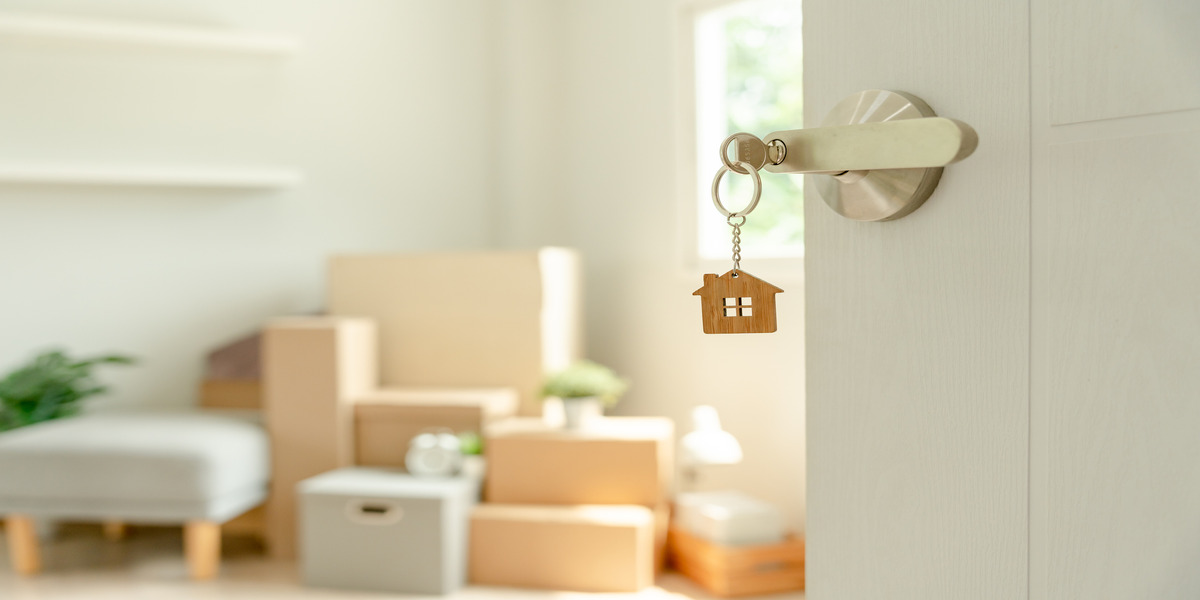A pair of house keys with a house-shaped keychain inserted into the lock of an open door, with a bright, airy room in the background featuring cardboard moving boxes and minimalist furniture, symbolizing new homeownership.