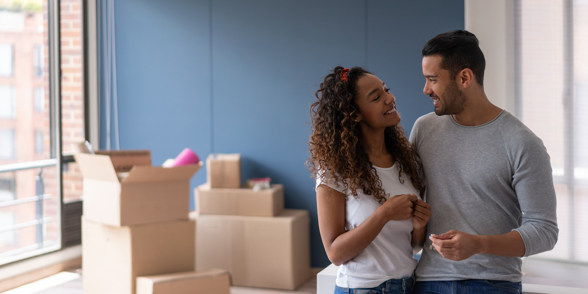 A smiling couple stands close together in a room with unpacked moving boxes, looking affectionately at each other, indicating a new chapter in their lives as homeowners.