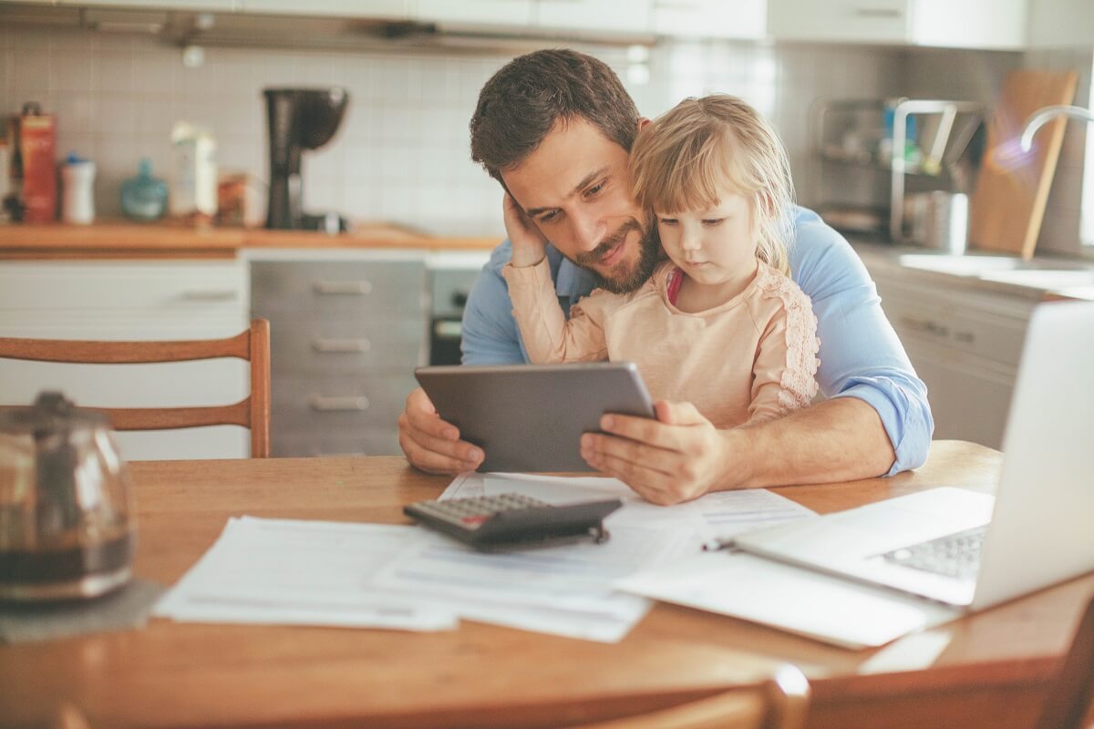 A father and toddler look at a tablet while doing paperwork at the kitchen table.