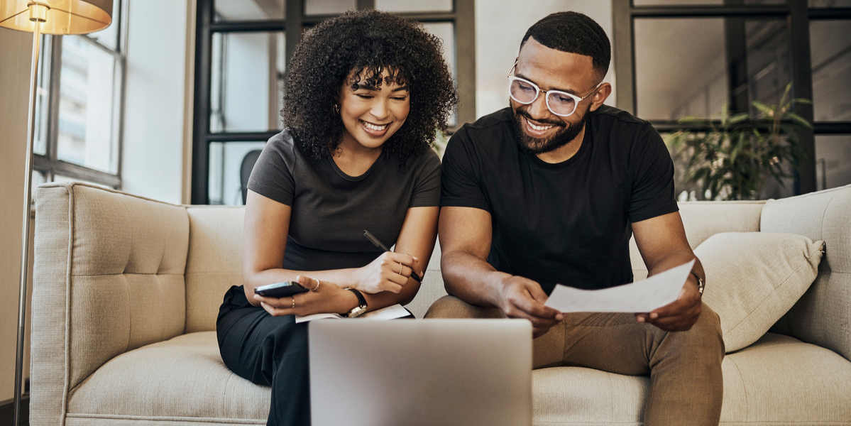 A man and woman are smiling and sitting together on a sofa with a laptop in front of them. She holds a smartphone and pen, ready to take notes, while he reviews a document, suggesting a collaborative work session in a home setting.