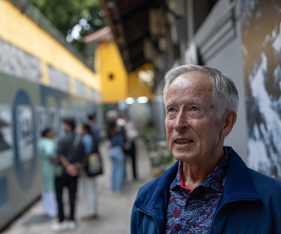 Robert "Bob" Jeffrey looking up at an outdoor photo exhibit in Vietnam, other visitors visible in the background.
