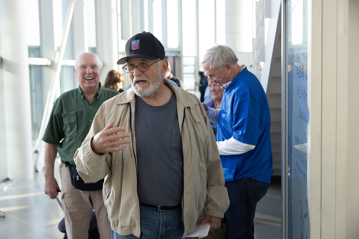 Dave Rodriguez talking with fellow veterans in an airport terminal, other group members visible and laughing in the background.