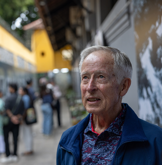 Robert "Bob" Jeffrey looking up at an outdoor photo exhibit in Vietnam, other visitors visible in the background.